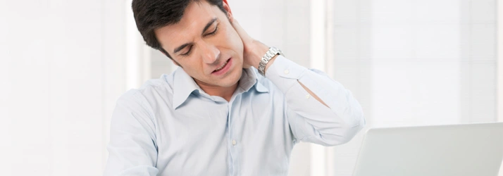 Individual at desk holding neck, suggesting neck strain or injury.